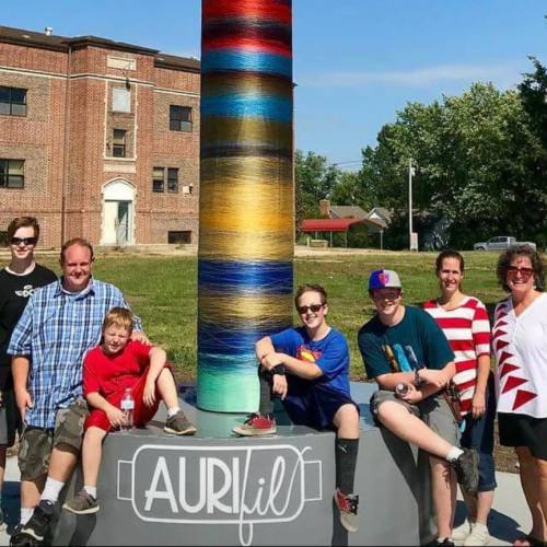 A group of seven people poses beside a colorful yarn sculpture on a sunny day, with a building in the background.