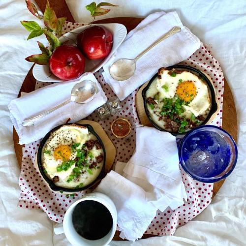 A wooden tray with two skillet dishes of eggs, herbs, and meat, surrounded by apples, utensils, and drinks on a white cloth.