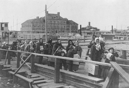 Black and white photo of a group of immigrants walking along a dock, carrying belongings, with buildings in the background.