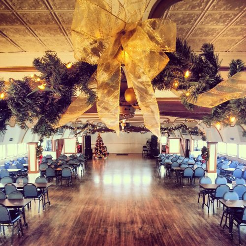 A festive hall decorated with garlands and lights, featuring a Christmas tree and rows of blue chairs.