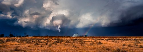 A dramatic landscape featuring dark storm clouds, a tornado, a rainbow, and a distant industrial building in a field.