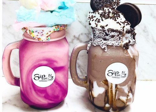 Two colorful milkshakes in mason jars: one topped with cotton candy and a figure, the other with whipped cream and cookies.