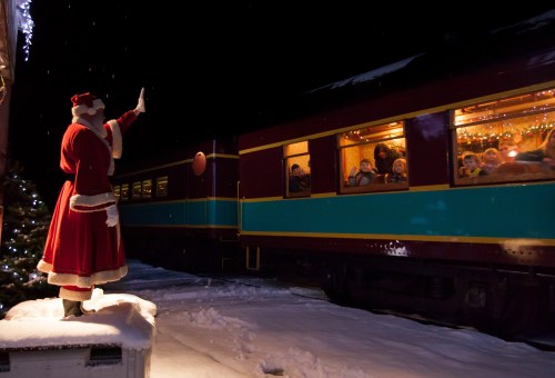 A Santa figure waves to children on a festive train, surrounded by snow and holiday lights at night.