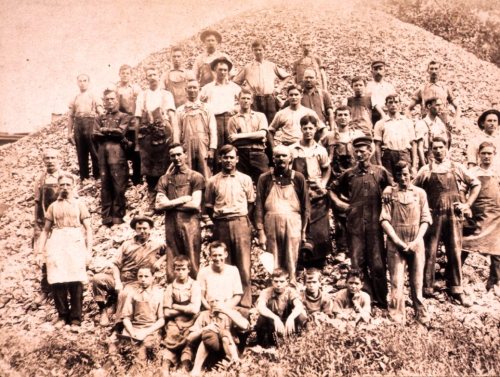 A large group of men and boys in work attire, posing in front of a pile of rocks, likely from the early 20th century.