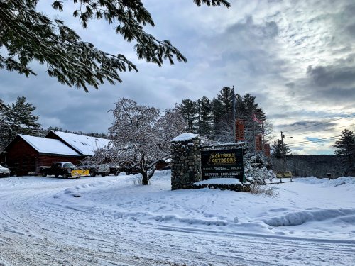 A snowy landscape featuring a rustic lodge and a sign for Northern Outdoors, surrounded by trees under a cloudy sky.