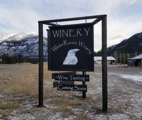 Sign for White Raven Winery with a wine tasting announcement, set against a snowy mountain backdrop.
