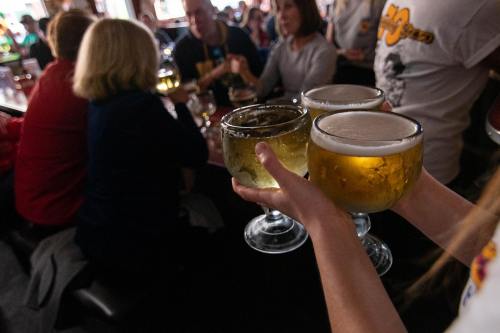 A person holds three large glasses of beer in a lively bar setting with people socializing in the background.