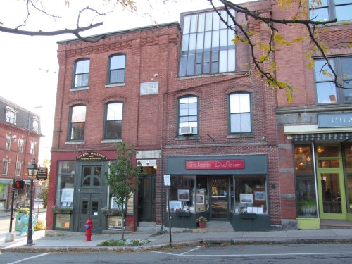A charming street view of brick buildings, featuring a gallery and a gelato shop, with autumn leaves in the foreground.