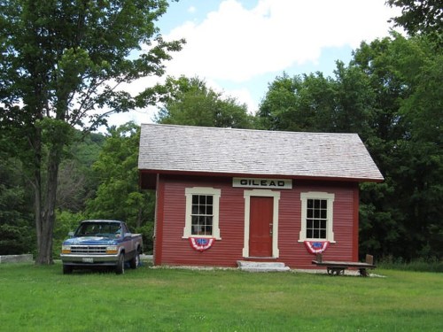 A red building labeled "Gilead" with a truck parked outside, surrounded by trees and grass under a blue sky.