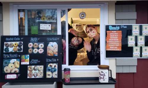 Two smiling employees in masks wave from a donut shop window, surrounded by a menu of assorted donuts and drinks.