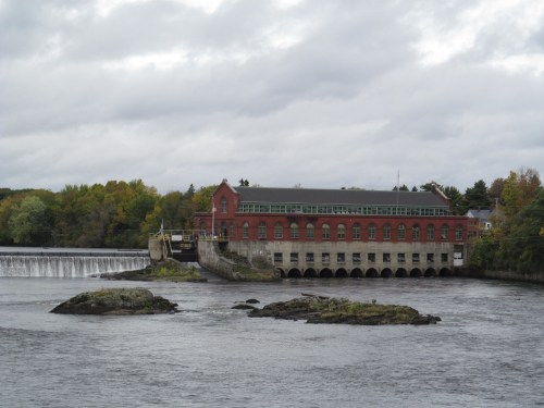 Historic red brick building by a river, with a dam and rocky outcrop, under a cloudy sky.