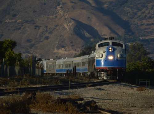 A blue and silver train travels along a track with mountains in the background, under clear skies.