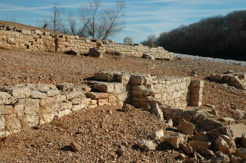 Ruins of stone walls on a rocky landscape, with sparse trees and a clear blue sky in the background.