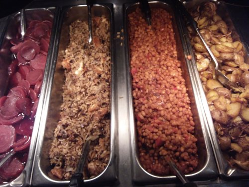 A buffet with four trays: sliced beets, rice with chicken, baked beans, and roasted potatoes, each with serving utensils.