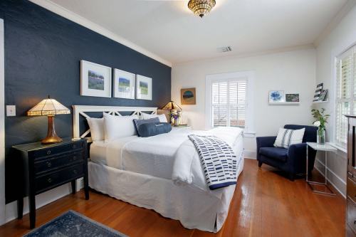 Cozy bedroom with a bed, nightstand, armchair, and artwork on the walls, featuring warm lighting and wooden floors.