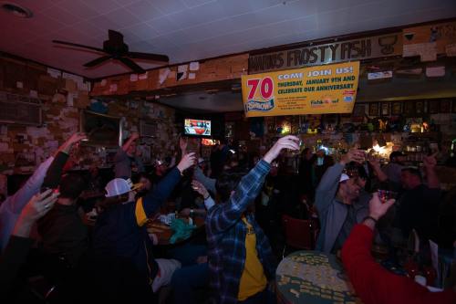 A lively bar scene with patrons raising glasses in celebration, surrounded by decorations and a banner for an anniversary event.
