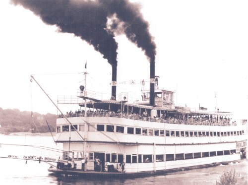 A historic steamboat with two smokestacks, filled with passengers, sailing on a river. Black and white photo.