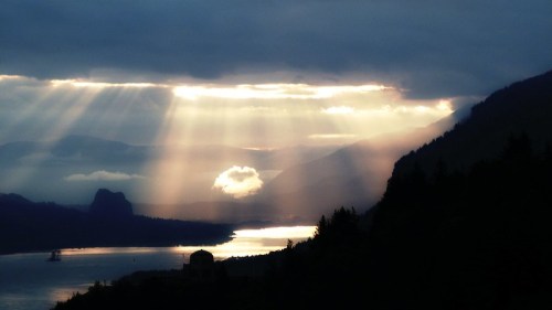 Sunlight streams through clouds over a river, surrounded by mountains and trees, creating a serene, dramatic landscape.