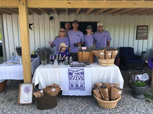 A group of six people in matching shirts at a lavender stand, with products displayed on a table under a wooden shelter.