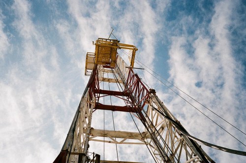 A tall drilling rig against a blue sky with scattered clouds, viewed from below.
