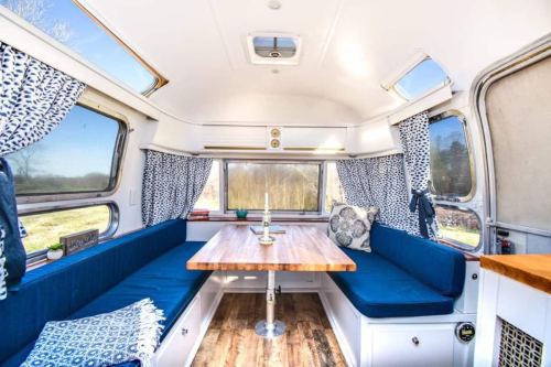 Cozy interior of a camper with a wooden table, blue seating, and large windows, surrounded by patterned curtains.