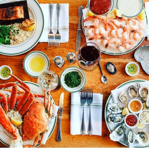 A table set with seafood dishes, including crab, shrimp, oysters, and sauces, surrounded by utensils and drinks.