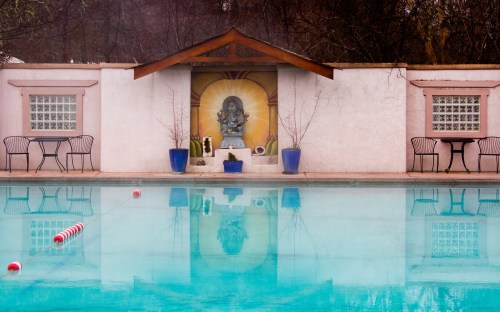 A serene pool area with a statue backdrop, surrounded by blue pots and chairs, reflecting in the calm water.