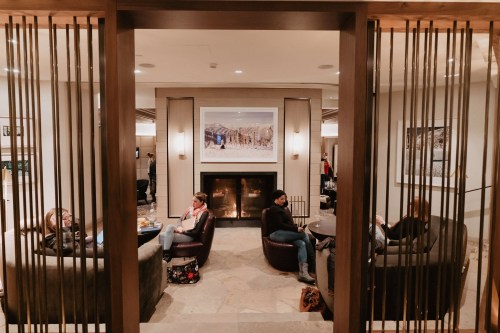 A cozy lounge area with people sitting in armchairs, a fireplace, and artwork on the wall, framed by wooden slats.