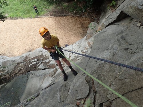 A child in a yellow shirt and helmet rappels down a rocky cliff, with a grassy area and another person in the background.