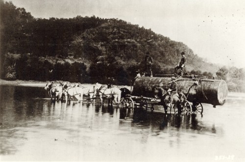 Two men stand on a large water tank pulled by horses crossing a river, surrounded by a hilly landscape.