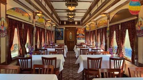 Interior of a vintage train dining car, featuring tables with white tablecloths, rich curtains, and decorative murals.
