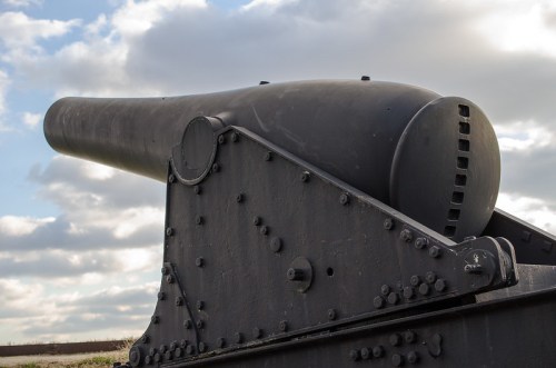 Close-up of a large black cannon against a cloudy sky, showcasing its barrel and mounting details.