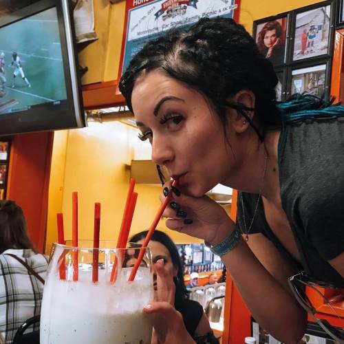 A woman with braided hair sips from a large drink with multiple straws in a vibrant restaurant setting.