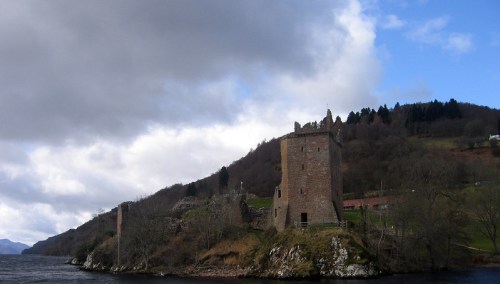A historic stone castle on a lakeshore, surrounded by trees and hills under a cloudy sky.