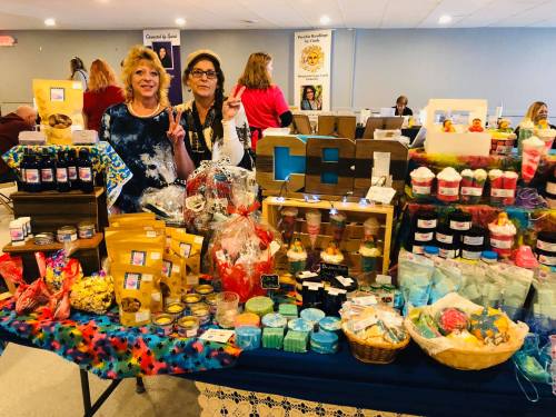 Two women pose with peace signs in front of a colorful display of handmade goods and crafts at a market event.