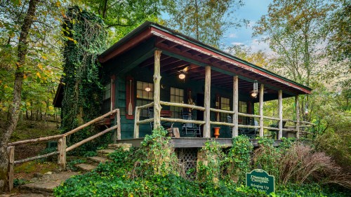 A cozy cabin surrounded by greenery, featuring a porch with rocking chairs and rustic wooden details.