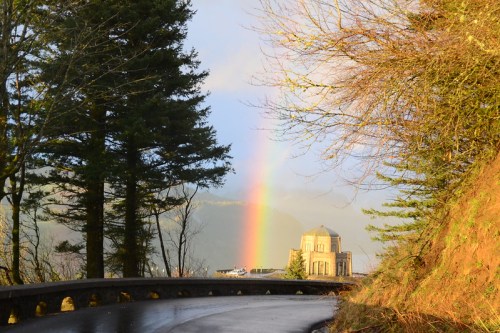 A winding road leads to a building with a dome, framed by trees and a vibrant rainbow in the background.