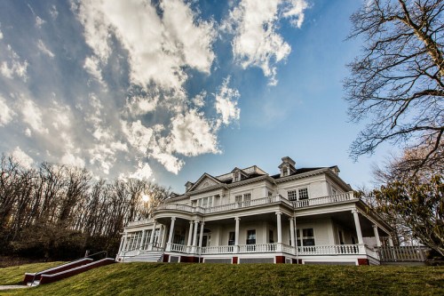 A large, historic white mansion with a wraparound porch, set against a blue sky and wispy clouds.