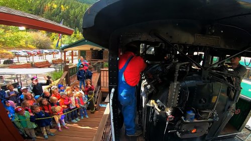 A train conductor interacts with a group of excited children at a scenic train station surrounded by trees.
