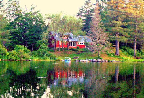 A serene lakeside scene featuring a red house surrounded by trees, reflecting in the calm water.