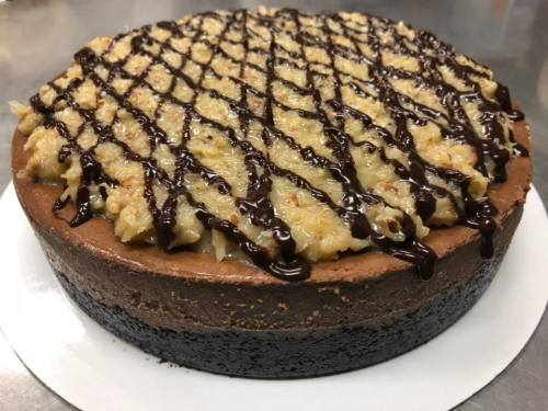 A rich chocolate cheesecake topped with coconut and drizzled with chocolate sauce, on a white plate.