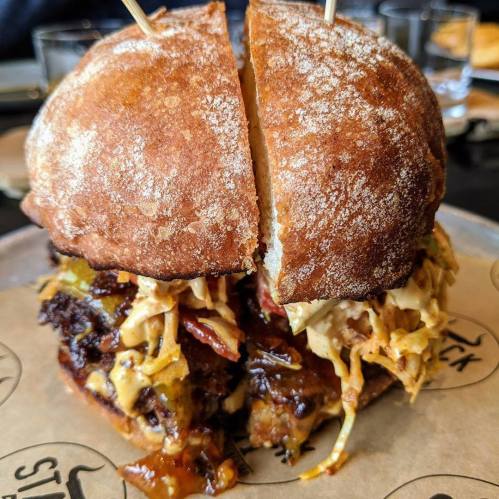 A close-up of a split sandwich with pulled meat, coleslaw, and pickles on a rustic bun, served on parchment paper.