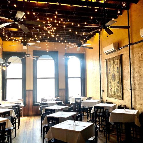 A cozy restaurant interior with tables set for dining, string lights overhead, and large windows letting in natural light.