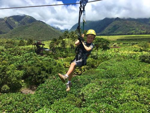 A child in a helmet zip-lining over lush green trees with mountains and clouds in the background.
