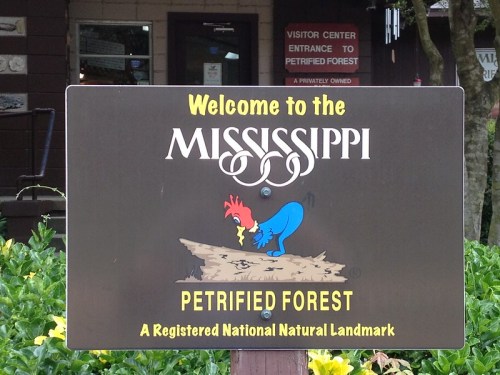 Sign welcoming visitors to the Mississippi Petrified Forest, featuring a cartoon rooster and natural landmark designation.