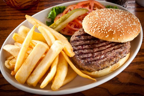 A sesame seed bun with a grilled burger patty, served with lettuce, tomato, pickles, and a side of golden fries.