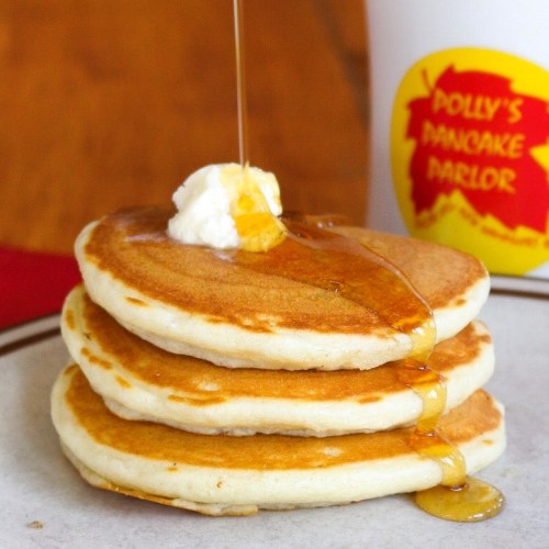 Three fluffy pancakes stacked on a plate, topped with butter and drizzled with syrup, next to a cup from Polly's Pancake Parlor.