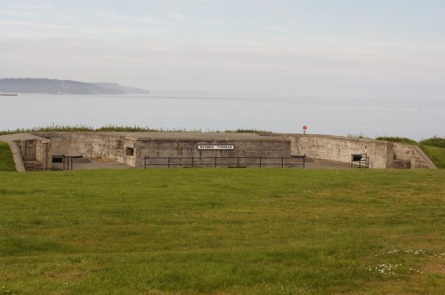 A concrete military fortification overlooking a calm sea, surrounded by green grass and distant hills.