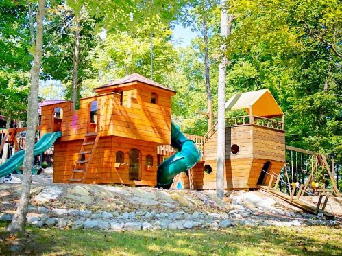 A wooden playground with slides, climbing structures, and a treehouse, surrounded by trees and greenery.