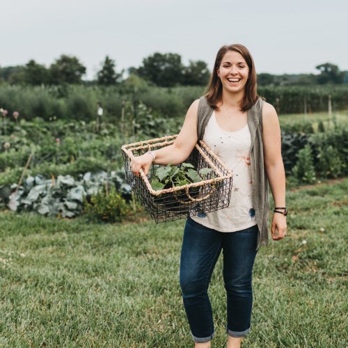 A smiling woman holds a basket of vegetables in a lush garden, surrounded by greenery and crops.